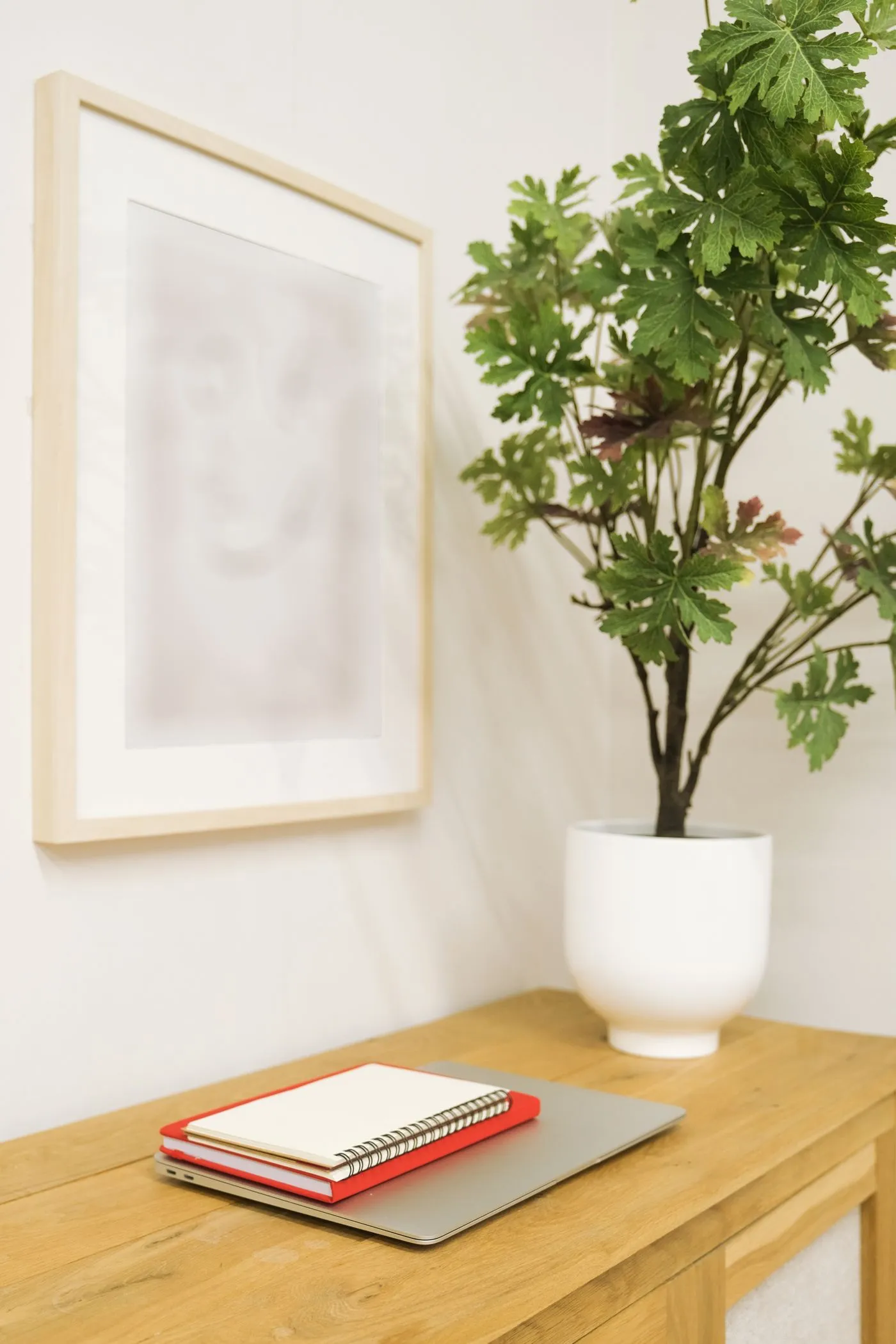 木の机に置かれたノートとノートパソコン、壁の額と観葉植物