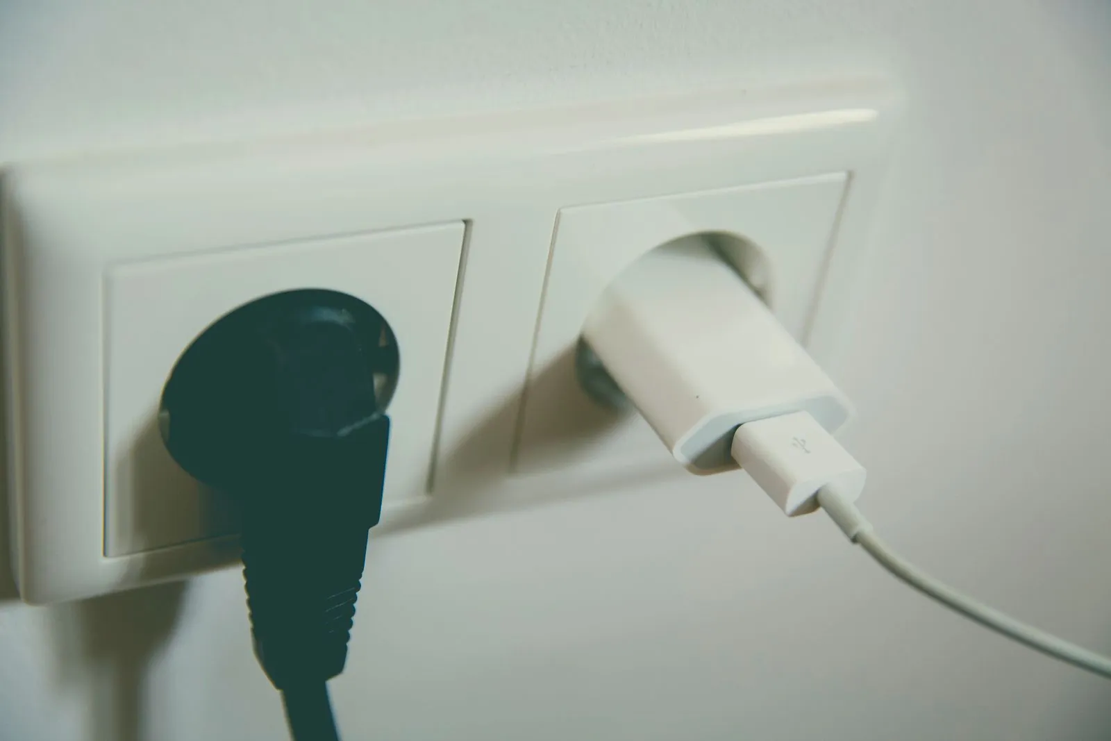 白い壁のコンセントに、黒いプラグと白い充電ケーブルが並んで差し込まれている