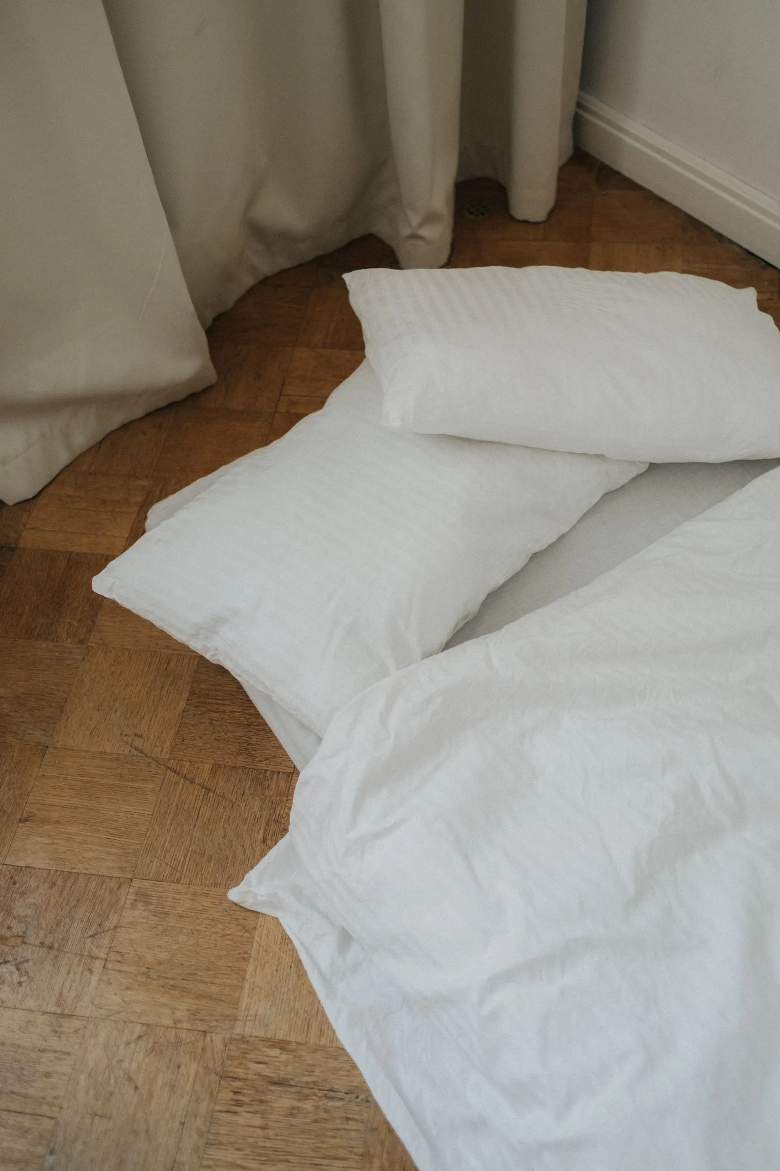 カーテンの前の木の床に置かれた白い枕2つと、その先に続く白い掛け布団