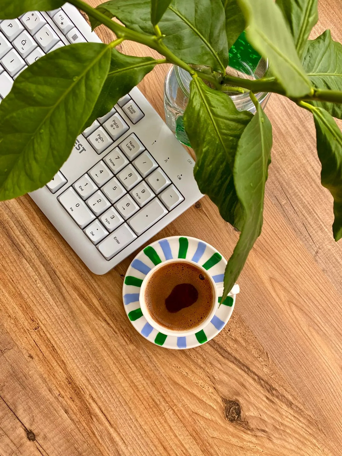 木目の机の上で、白いキーボードのテンキー部分の脇に緑の葉と湯気の立つコーヒーが置かれている