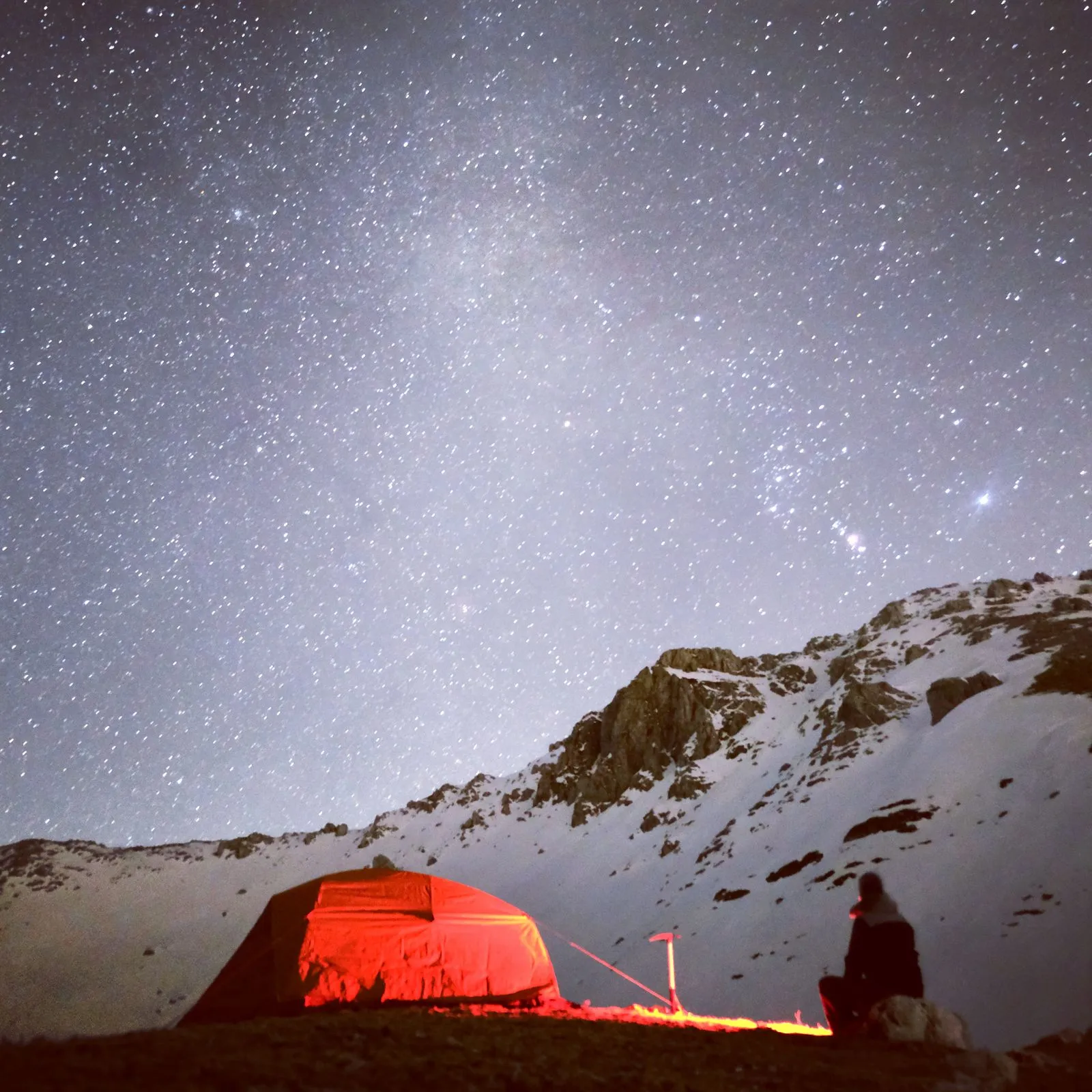 雪山の夜、星空の下で赤く光るテントと座る人物のシルエット