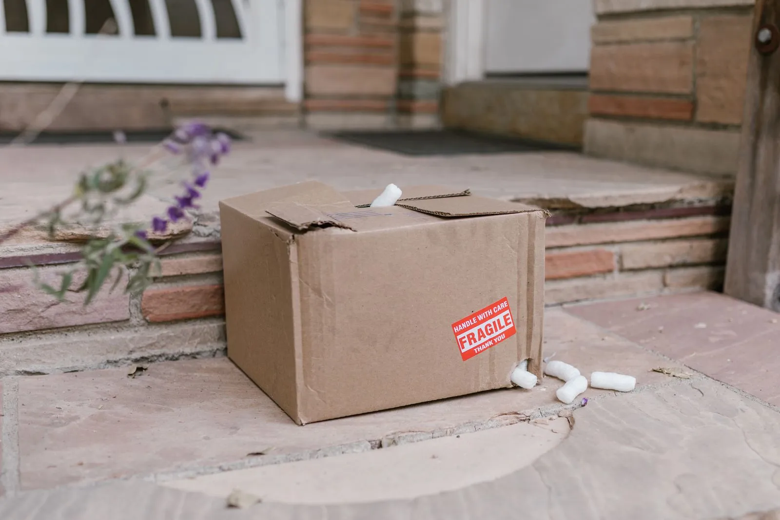 玄関ポーチの段差に置かれた段ボール箱。「FRAGILE」のシールが貼られ、白い緩衝材が周りに散らばっている