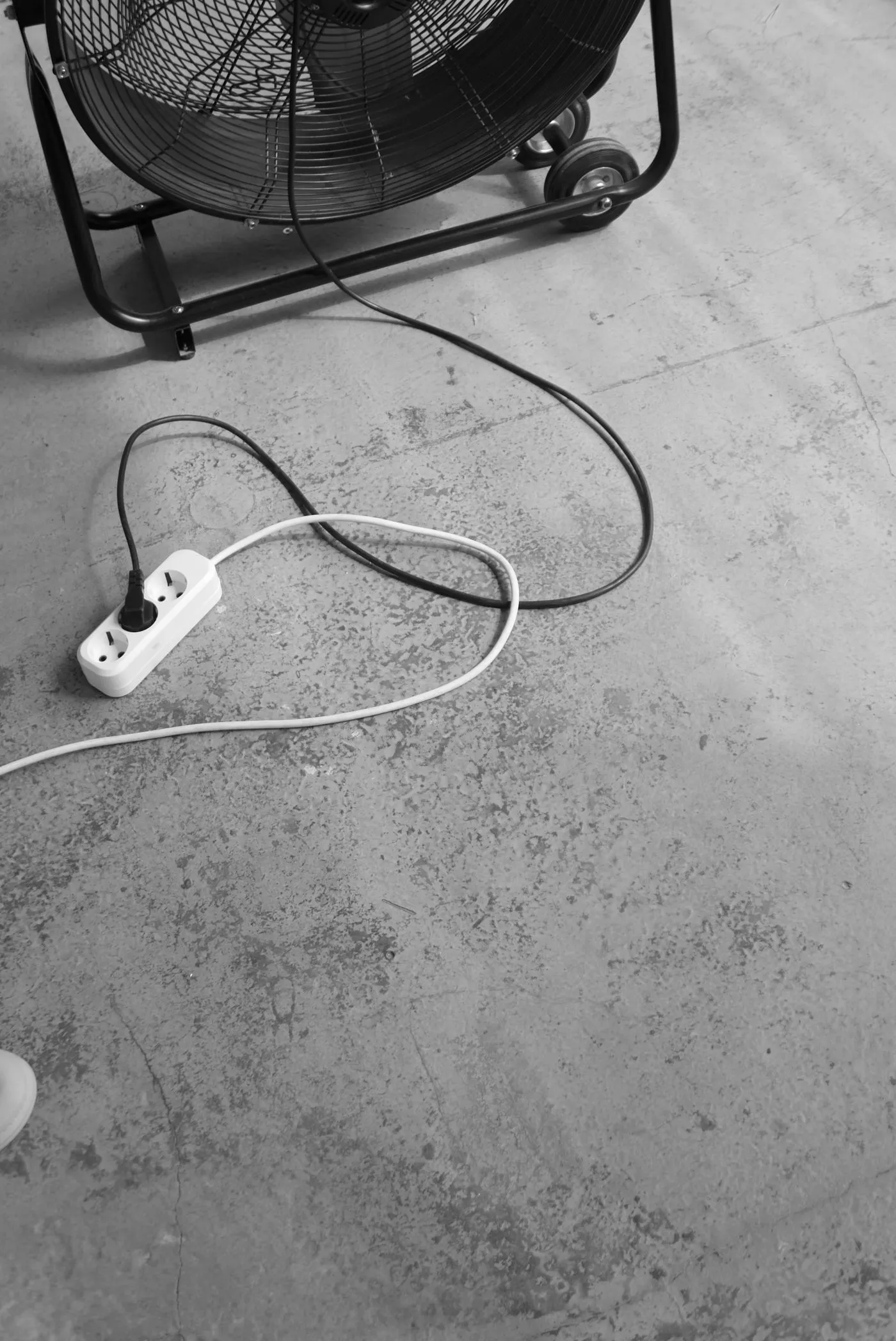 床に置かれた大型の送風機から伸びたコードが、電源タップにつながっている（白黒写真）